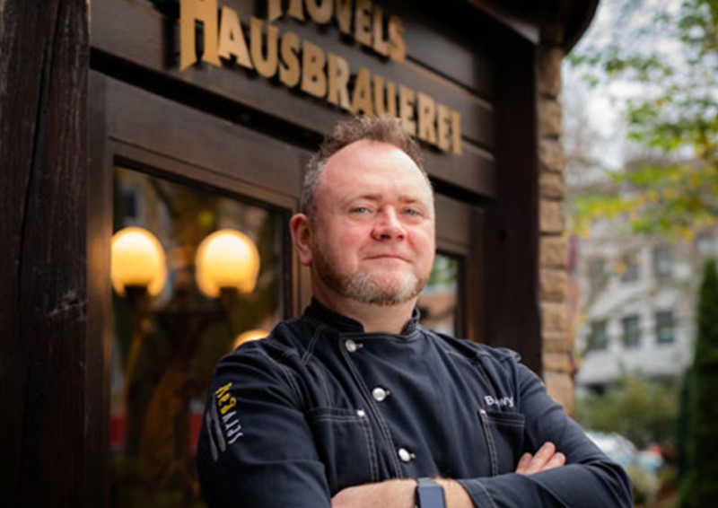 Ein Mann steht lächelnd mit verschränkten Armen vor einer Hausbrauerei.