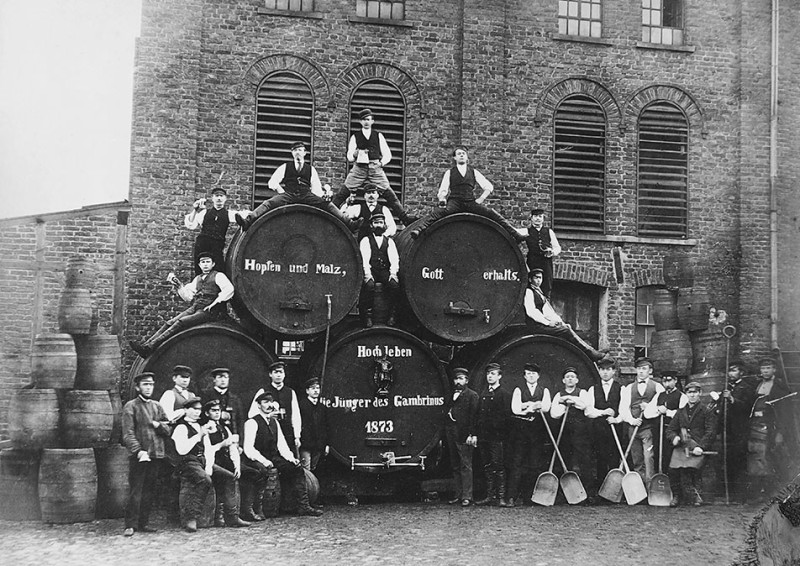 Eine Gruppe von Arbeitern posiert um 1873 vor großen Bierfässern vor einer Backsteinmauer.