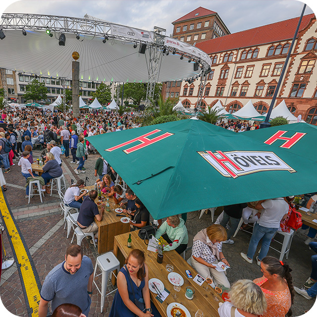 Menschen essen und unterhalten sich an Tischen unter Sonnenschirmen bei einem Stadtfest.