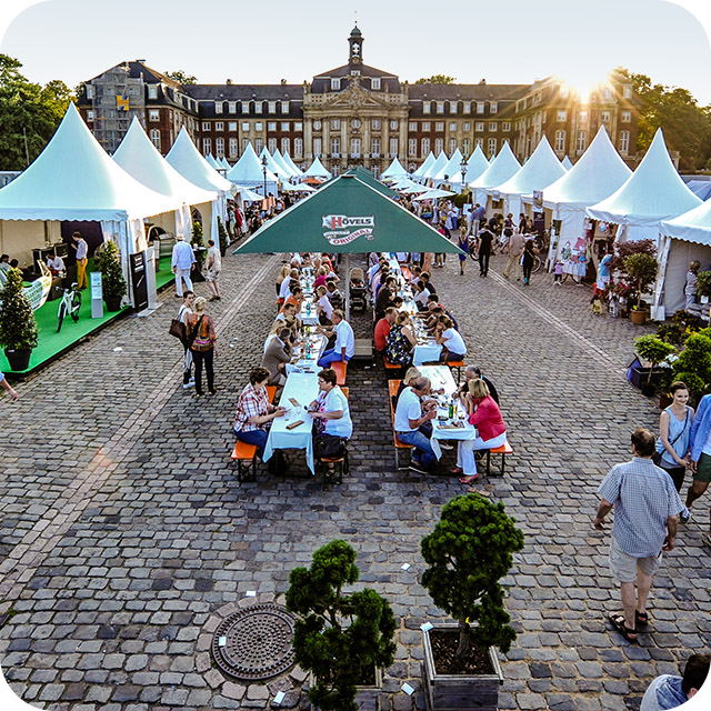 Menschen essen und unterhalten sich an Tischen bei einem Freiluft-Event vor einem großen historischen Gebäude.