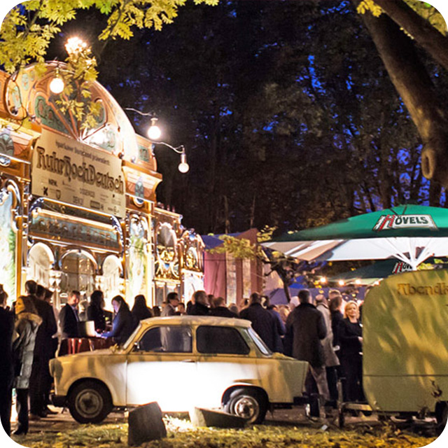 Ein belebter Abendmarkt mit Personen, die sich um einen illuminierten Stand und ein Auto versammeln.