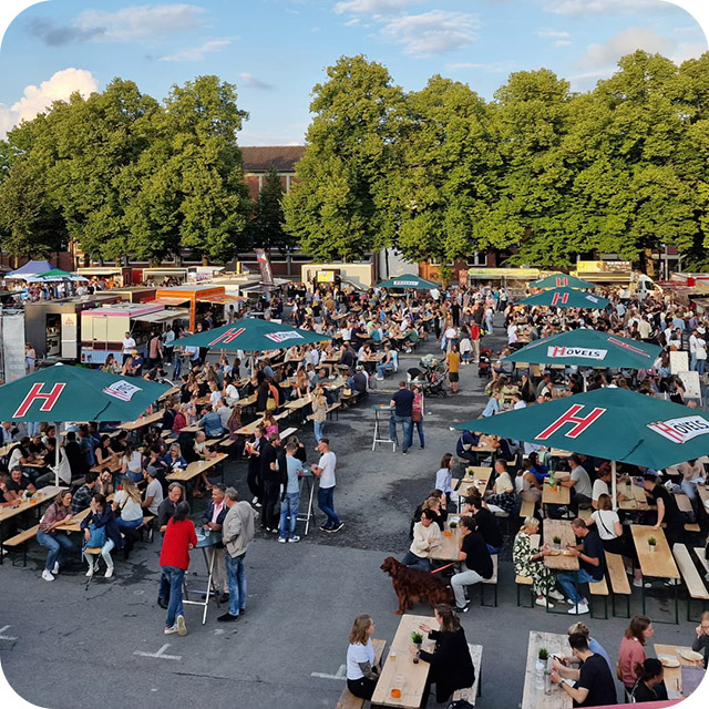 Viele Menschen sitzen an Tischen bei einem Freiluftmarkt mit Essensständen.
