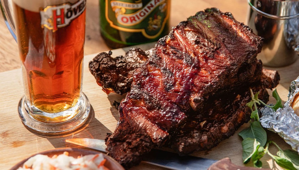 Auf einem Holzbrett liegt ein großes Stück gegrillte Rippe neben einem Glas Bier.