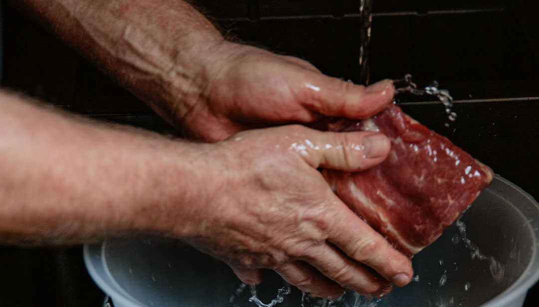 Zwei Hände waschen ein Stück rohes Fleisch unter fließendem Wasser.