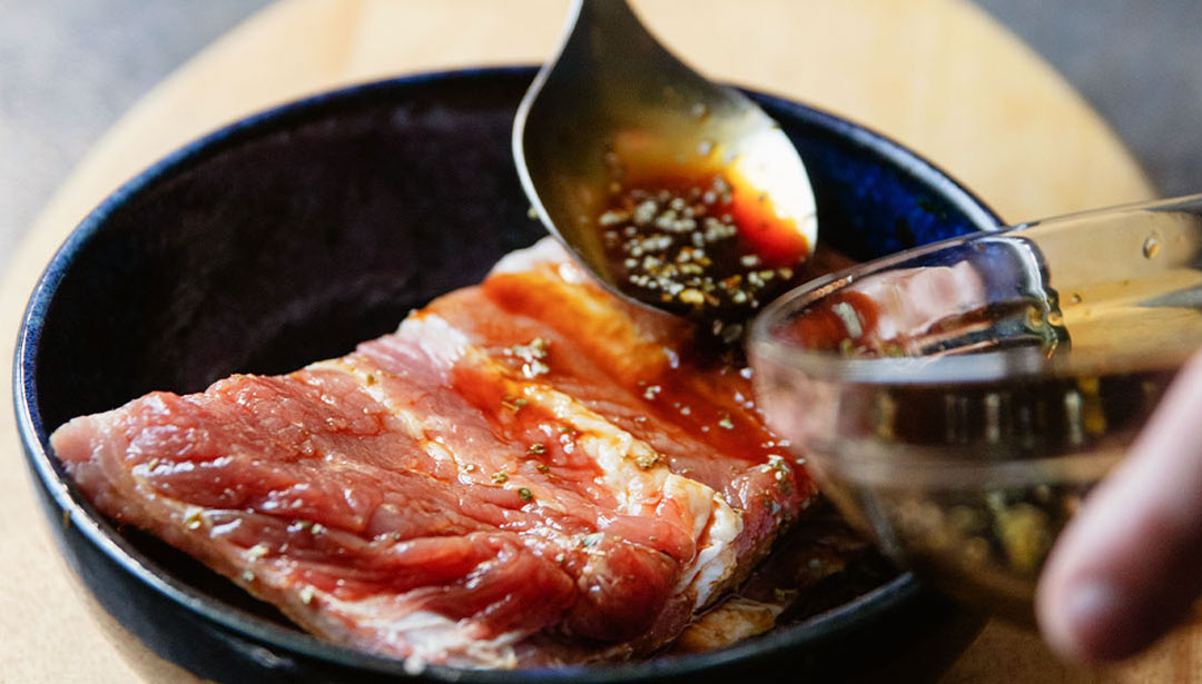 Eine Person gibt Sauce über rohes, gewürztes Fleisch in einer schwarzen Schüssel.