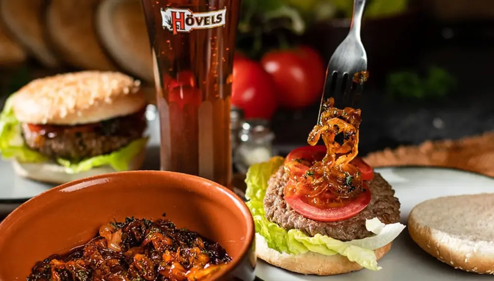 Ein offener Burger mit Salat und Tomate wird belegt, daneben ein Bierglas und ein zweiter Burger.