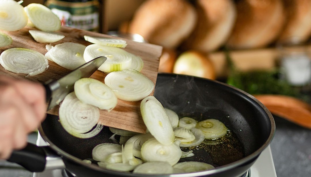 Eine Hand schneidet Zwiebeln, die auf einem Holzbrett liegen und in eine erhitzte Pfanne fallen.