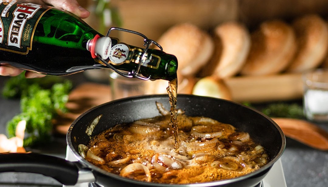 Eine Person gießt Bier in eine Pfanne mit bratenden Zwiebeln auf einem Herd.
