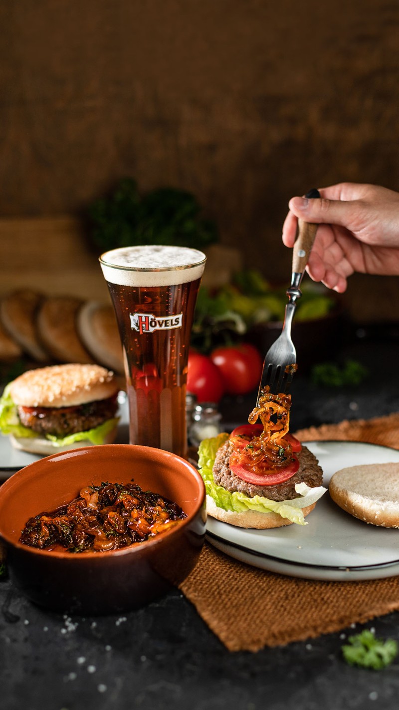 Ein Glas Bier steht neben einem Teller mit Hamburger und Beilagen. Eine Hand fügt ein Topping hinzu.