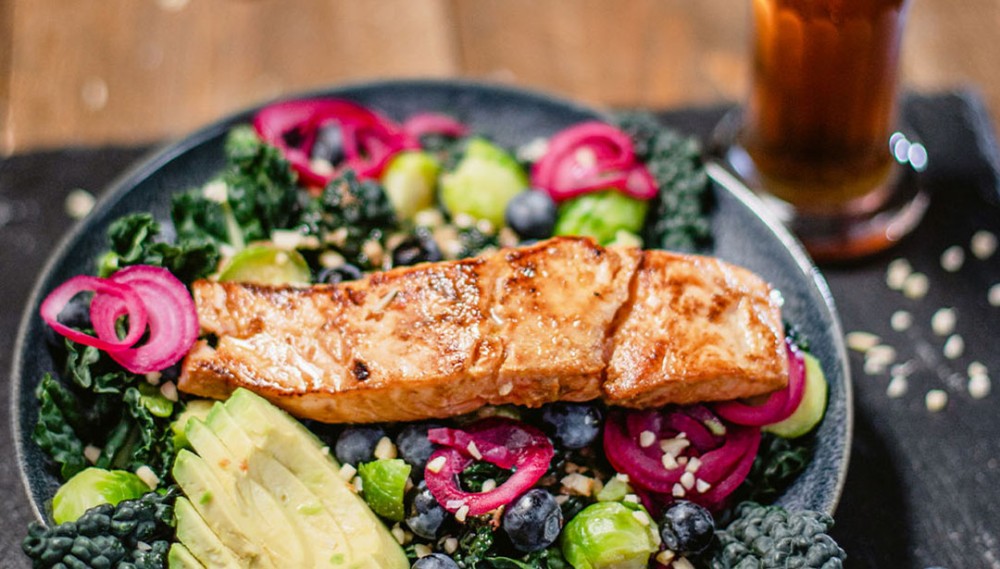 Ein gebratenes Lachsfilet liegt auf einem Bett aus Grünkohl und Avocadoscheiben, garniert mit roten Zwiebeln und Blaubeeren.