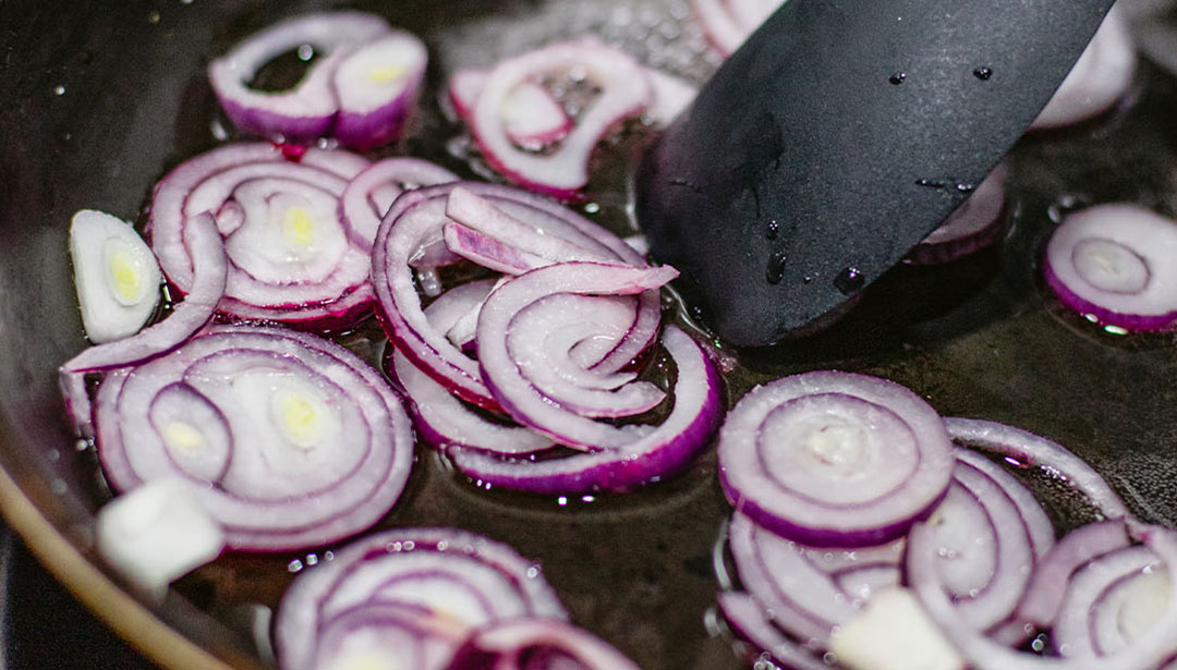 Eine Pfanne enthält gebratene rote Zwiebelringe, die von einer schwarzen Zange gehalten werden.