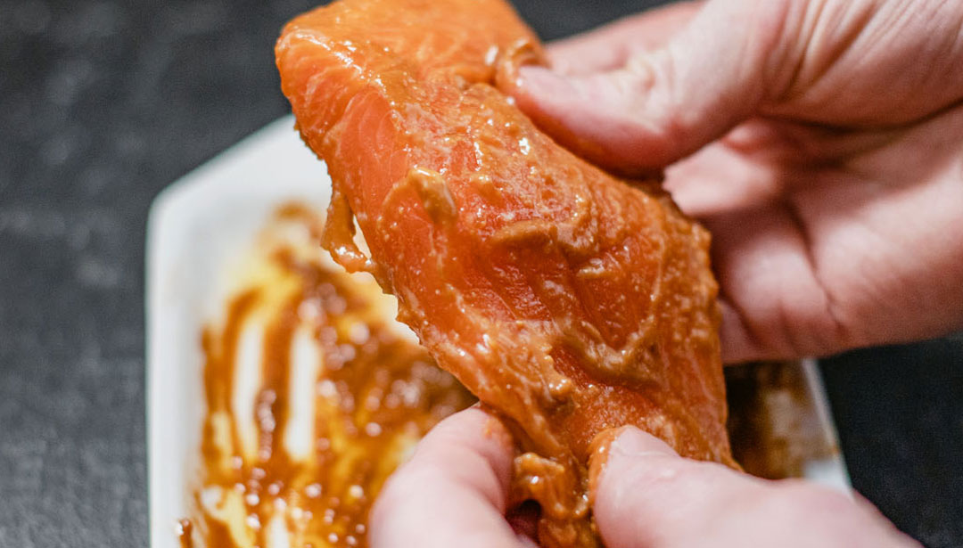 Eine Hand hält ein Stück marinierten Lachs über einer Schale.