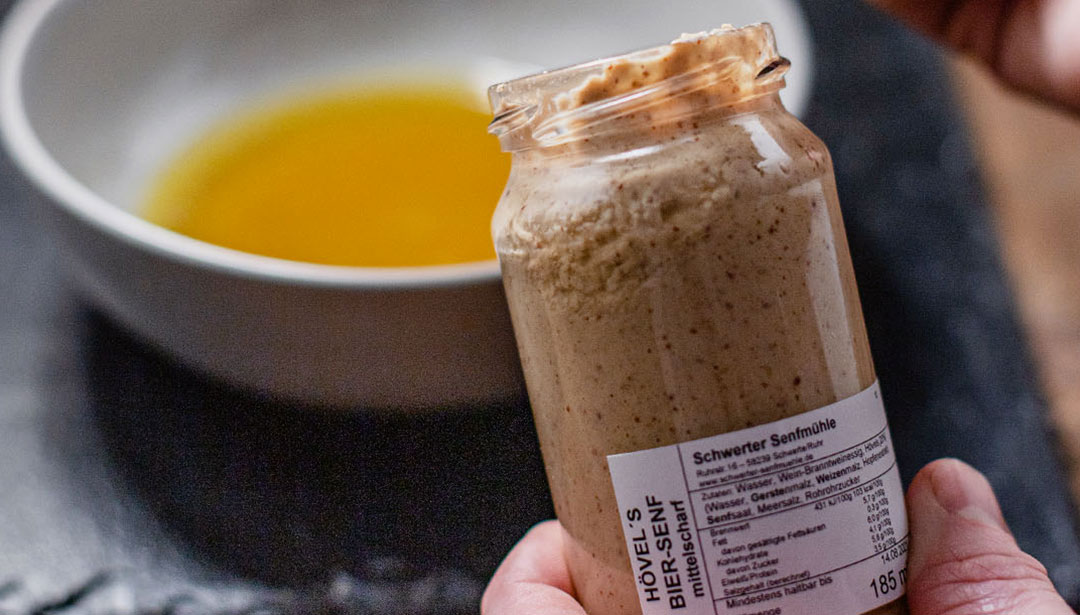 Ein Glas grobkörniger Senf wird neben einer Schale mit Öl gehalten.