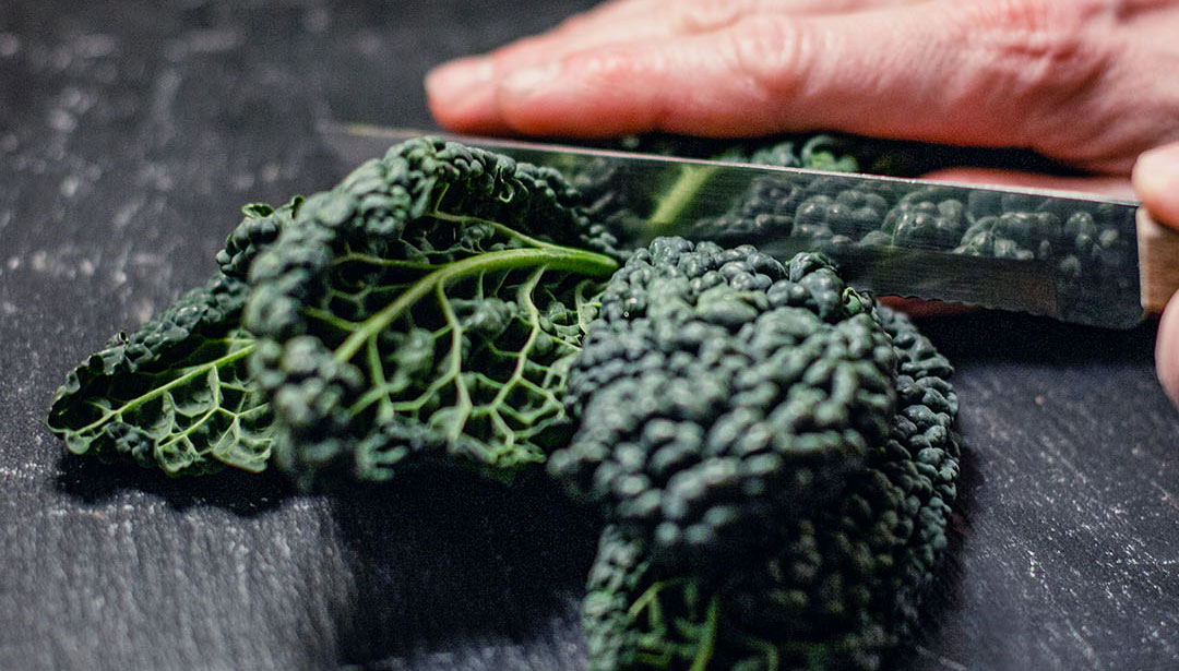 Eine Hand schneidet mit einem Messer Grünkohl auf einer dunklen Oberfläche.