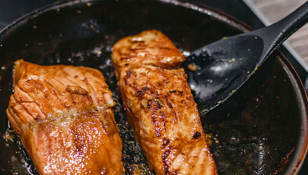 Zwei Lachsfilets braten in einer Pfanne und werden mit einem schwarzen Pfannenwender gewendet.