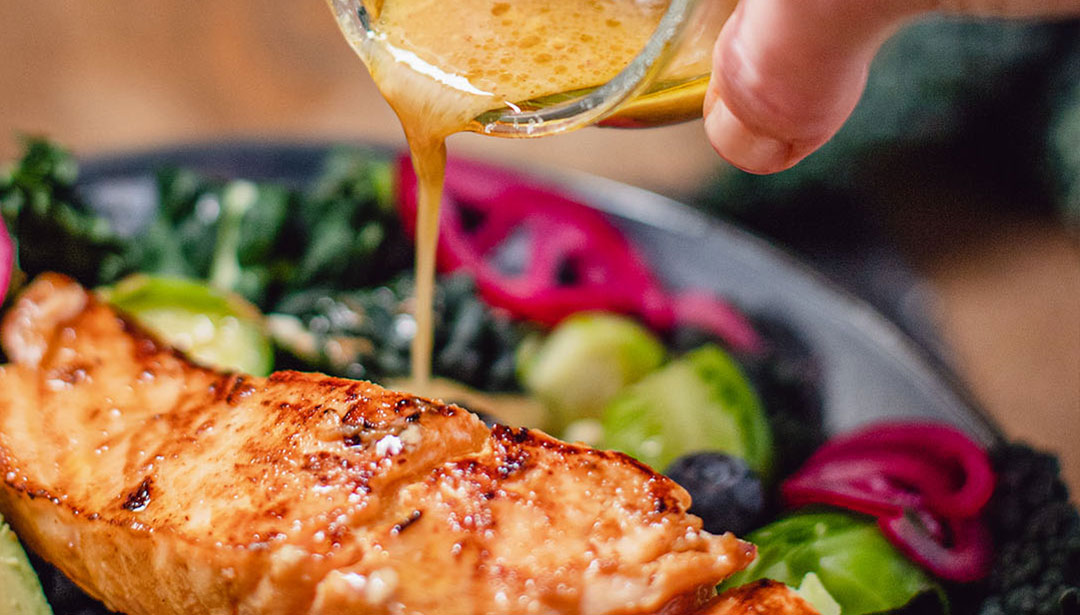 Eine Hand gießt Sauce über ein gebratenes Lachsfilet auf einem bunten Salatteller.