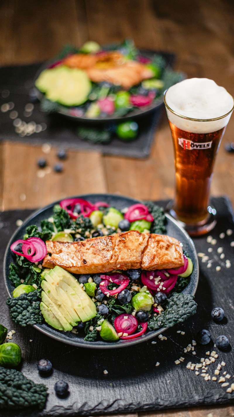 Ein gebratener Lachs liegt auf einem Salat mit Avocado, Rosenkohl und Beeren, daneben steht ein volles Bierglas.