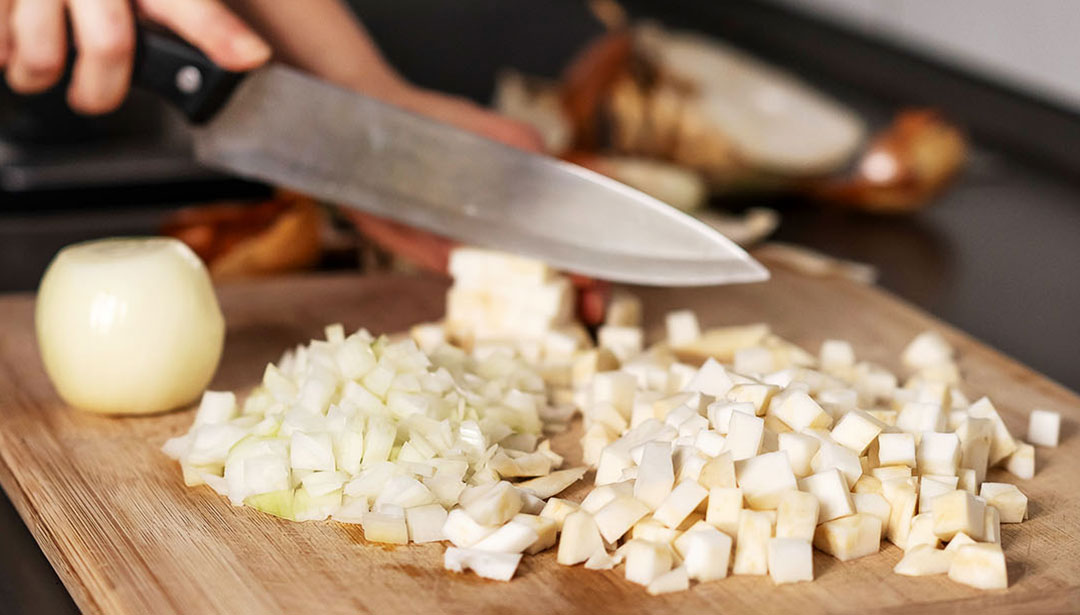 Eine Hand schneidet eine Zwiebel auf einem Holzbrett in Würfel.
