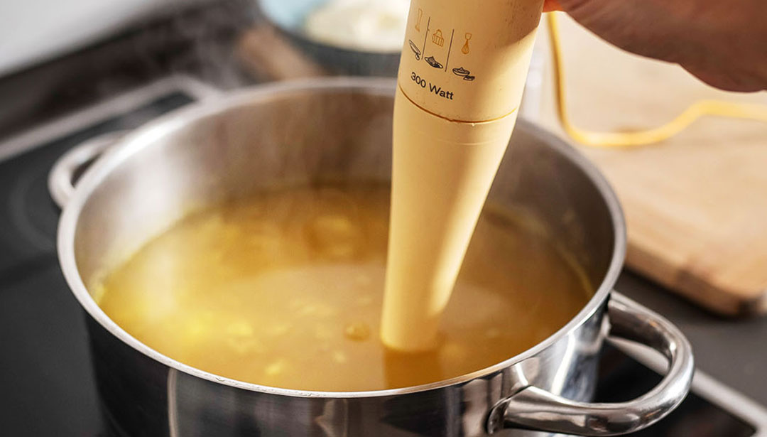 Ein Handmixer wird verwendet, um Suppe in einem Topf auf einem Herd zu pürieren.