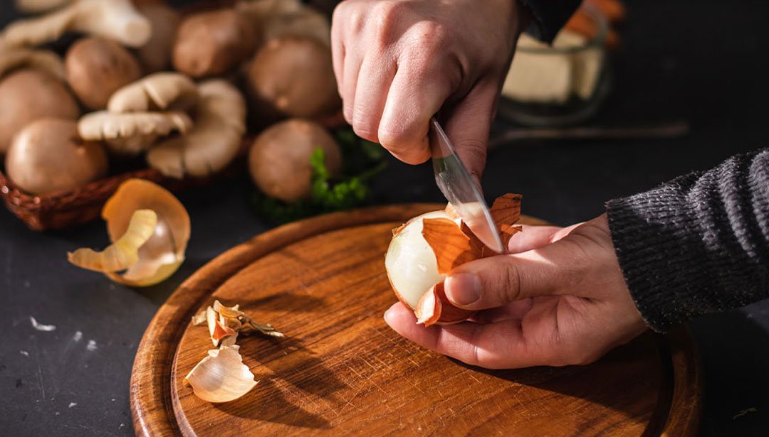 Eine Person schält eine Zwiebel auf einem Holzbrett, umgeben von Pilzen.
