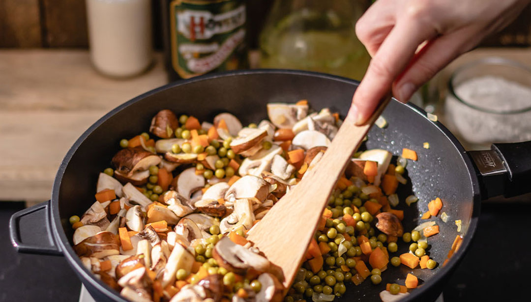 Eine Hand rührt Gemüse und Pilze in einer Pfanne auf einem Holztisch.