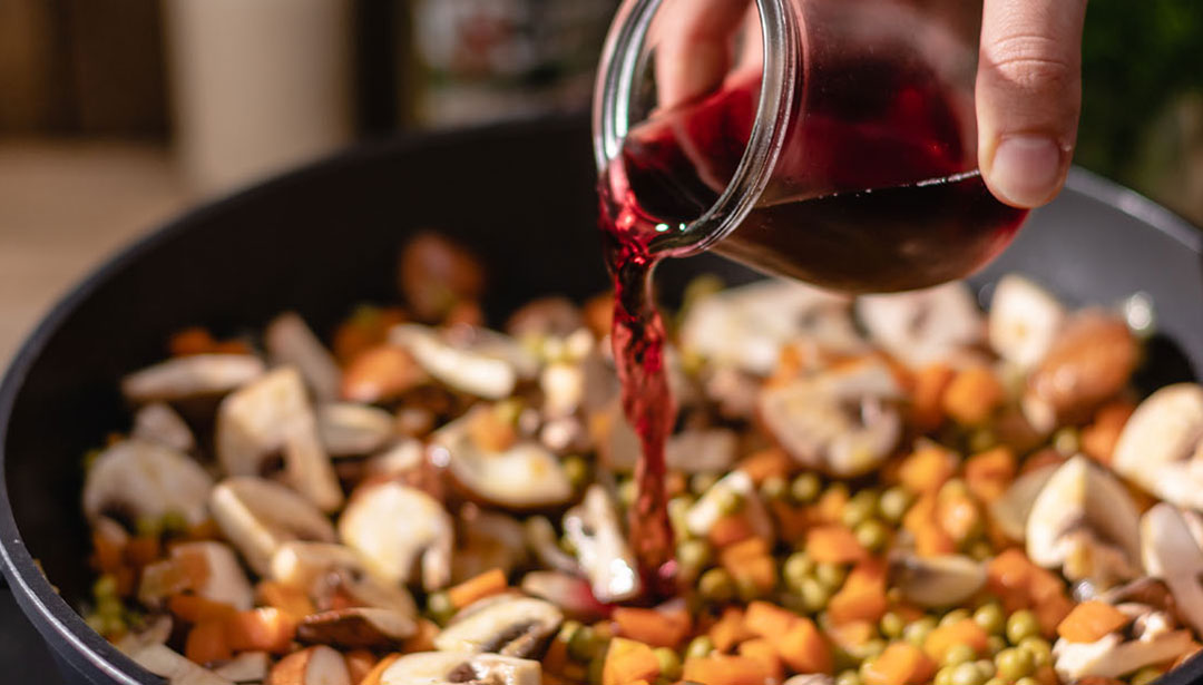 Eine Hand gießt rote Flüssigkeit in einen Topf mit geschnittenem Gemüse.