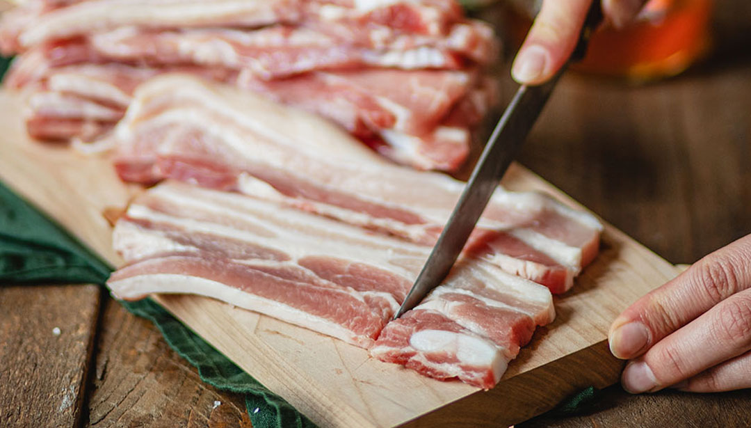 Eine Hand schneidet rohen Speck auf einem Holzbrett in Scheiben.