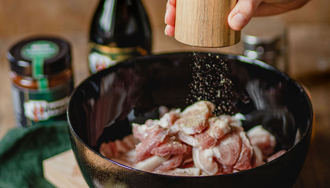 Eine Hand streut Pfeffer auf rohes Fleisch in einer schwarzen Schüssel.