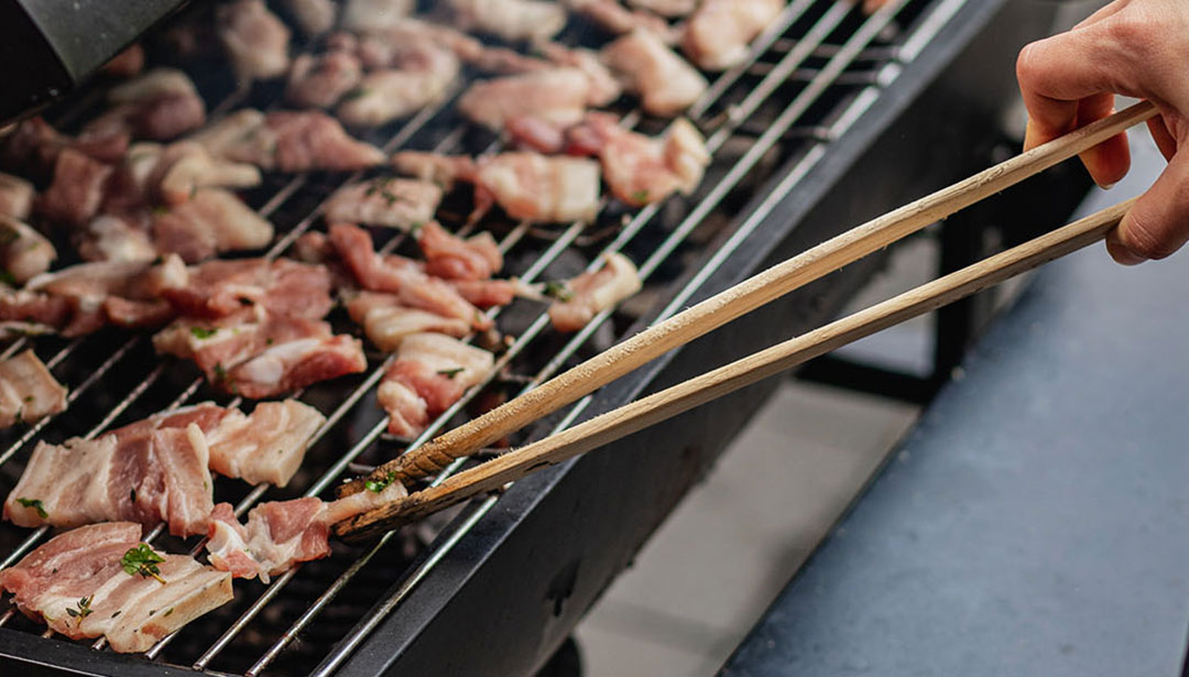 Eine Hand verwendet Zangen, um Fleischstücke auf einem Grill zu wenden.