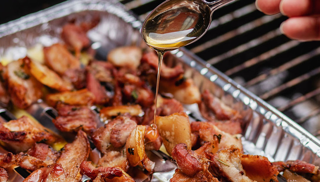 Gebratenes Fleisch und Kartoffeln werden auf einem Grill gegart, Öl wird gerade darüber geträufelt.