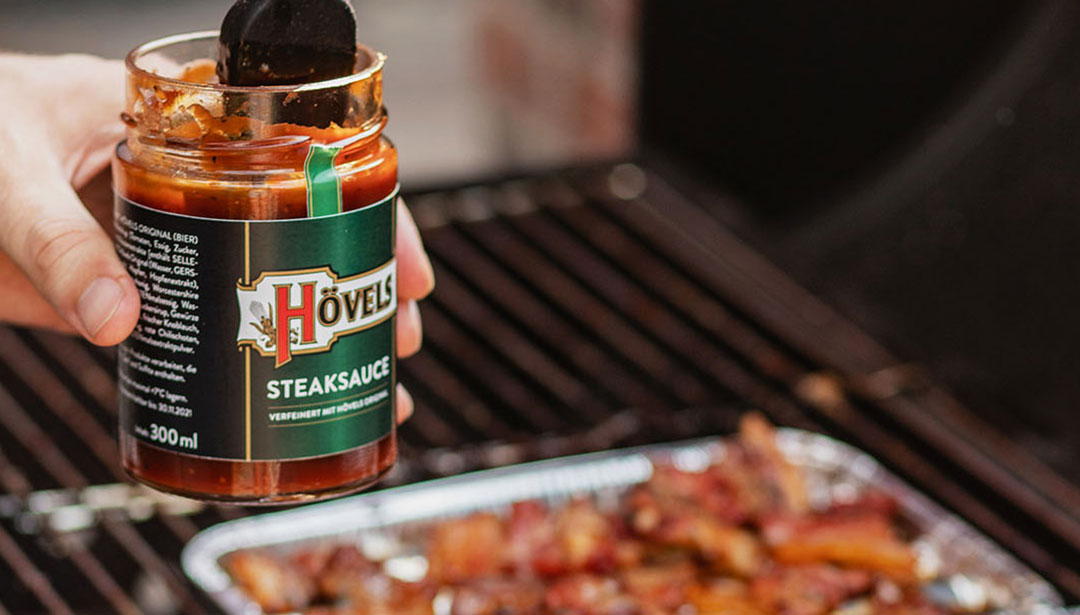 Eine Hand hält ein geöffnetes Glas mit Grillsauce über einer Aluschale mit Fleisch auf einem Grill.