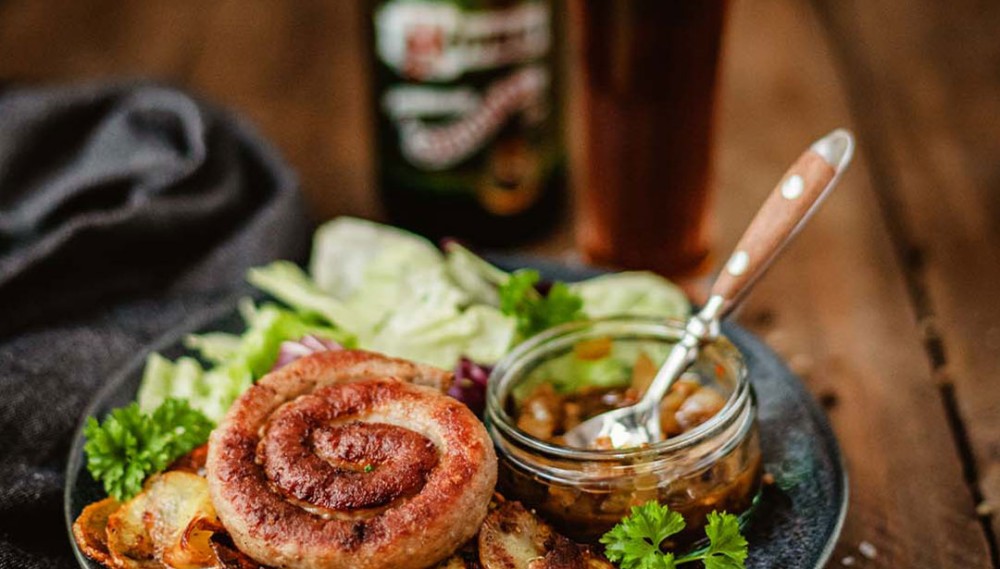 Ein gegrilltes Wurstspirale liegt auf einem Teller neben Salat und gebratenen Kartoffeln. Neben dem Teller steht ein Glas Bier.