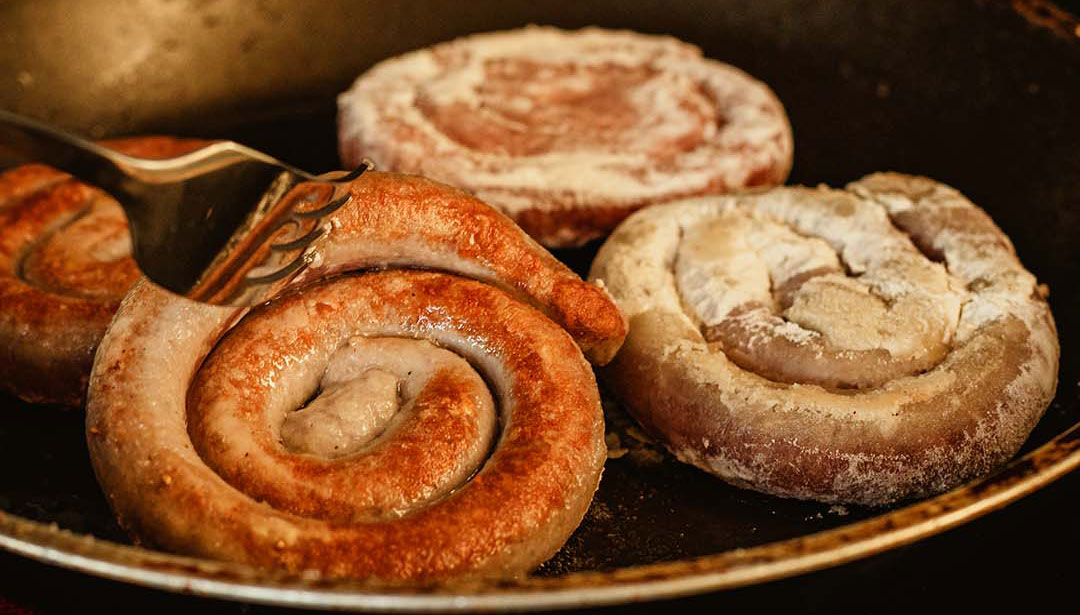 In einer Pfanne braten drei Wurstsorten, darunter eine spiralförmige Bratwurst.