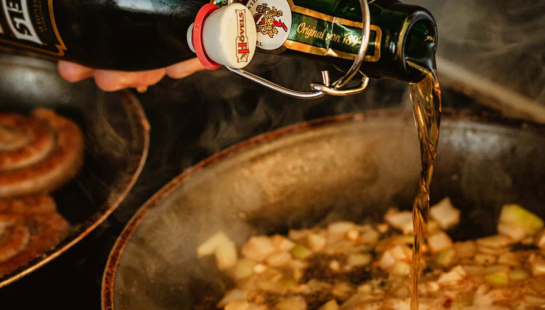 Eine Hand schüttet Flüssigkeit aus einer dunklen Flasche in eine Pfanne mit Bratgut.