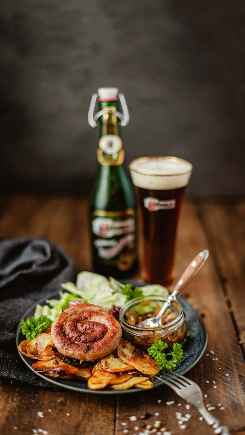 Ein Teller mit Bratwurst, Kartoffelscheiben und Salat neben einem Glas Bier steht auf einem Tisch.