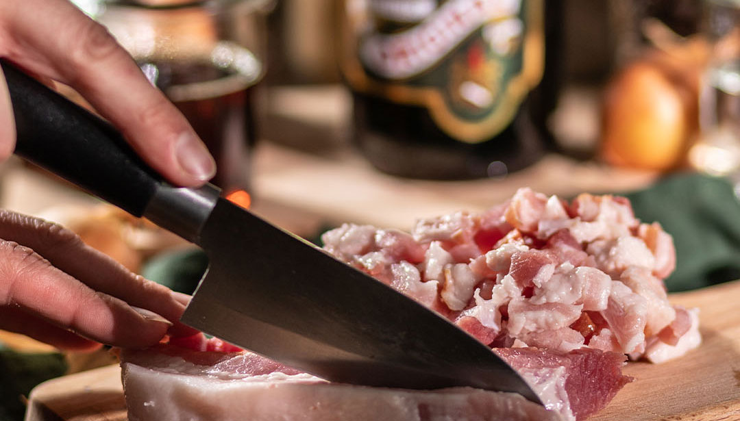 Eine Hand schneidet rohes Fleisch auf einem Holzbrett mit einem großen Messer.