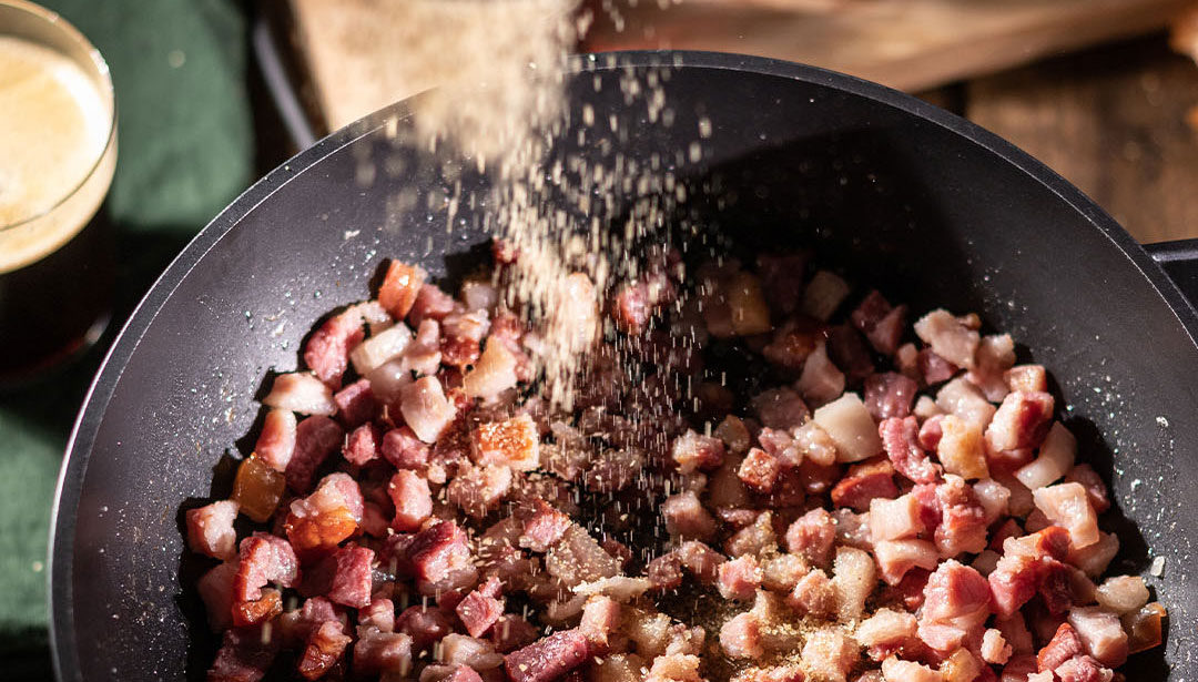 Gebratene Speckwürfel in einer Pfanne, darüber rieselt grobkörniges Salz.